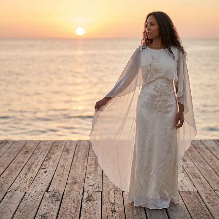 Elegant ivory satin column gown with intricate pearl floral embroidery and a flowing sheer chiffon cape, worn by a model standing on a wooden dock at sunset.