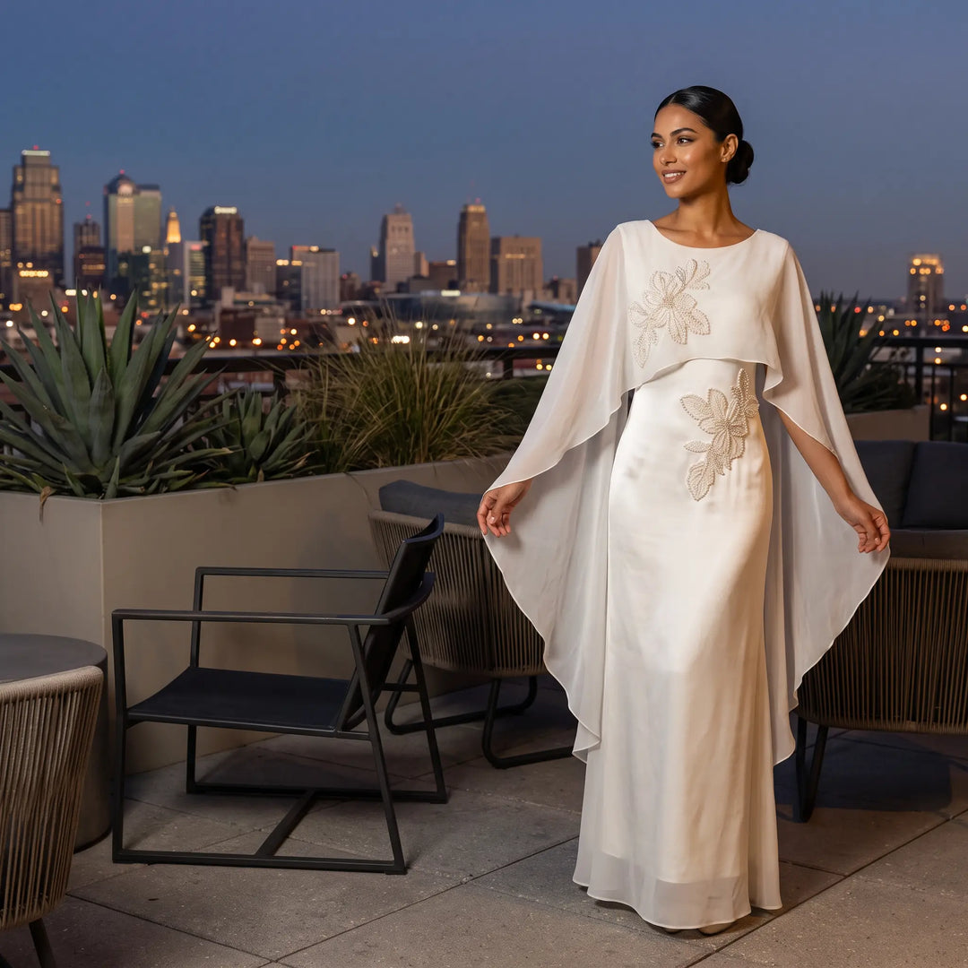Elegant ivory satin column gown with intricate pearl floral embroidery and a flowing sheer chiffon cape, modeled on a rooftop terrace at dusk with a city skyline view.