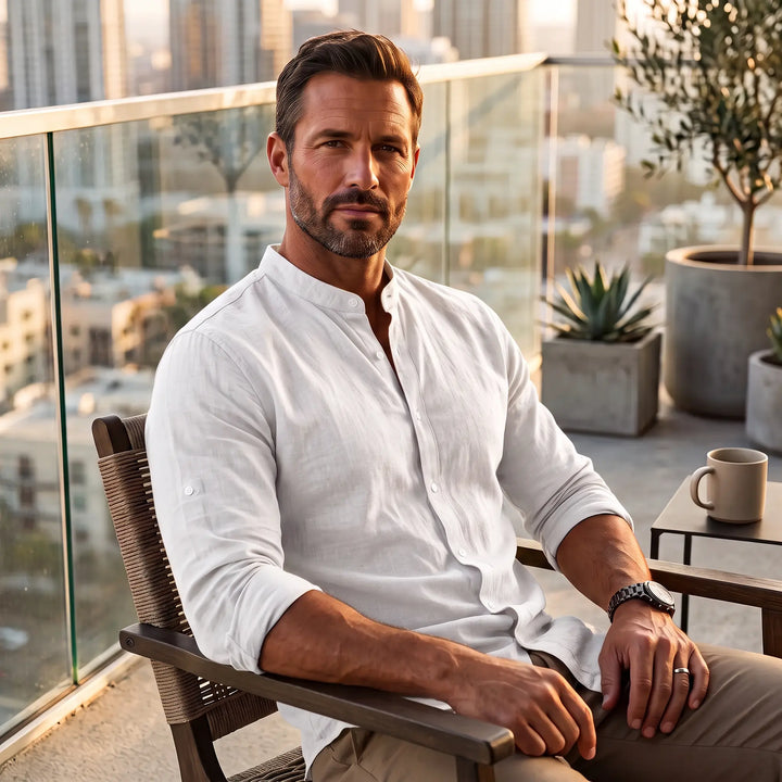 Man wearing a white Zenitho long-sleeve linen-blend shirt with a Mandarin collar, seated on a balcony overlooking a city skyline at sunset.