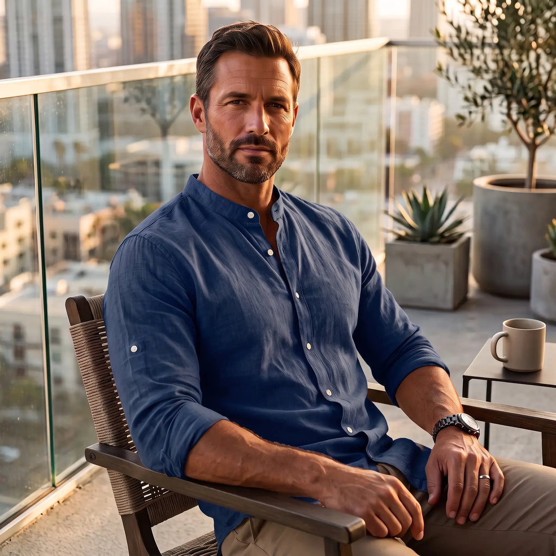 Man wearing a rich navy blue Zenitho long-sleeve linen-blend shirt with a Mandarin collar, seated on a balcony with a cityscape view, showcasing its relaxed fit and fabric texture.