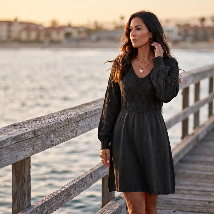 Model wearing a classic black V-neck knit sweater dress with a textured fish-scale pattern on the yoke and sleeves, cinched waist, and A-line skirt, standing on a wooden pier by the ocean.