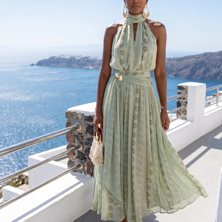 Woman wearing a mint green georgette maxi dress with a gold botanical print, halter neckline, keyhole cutout, and decorative buckle at the cinched waist, standing on a balcony overlooking the sea.