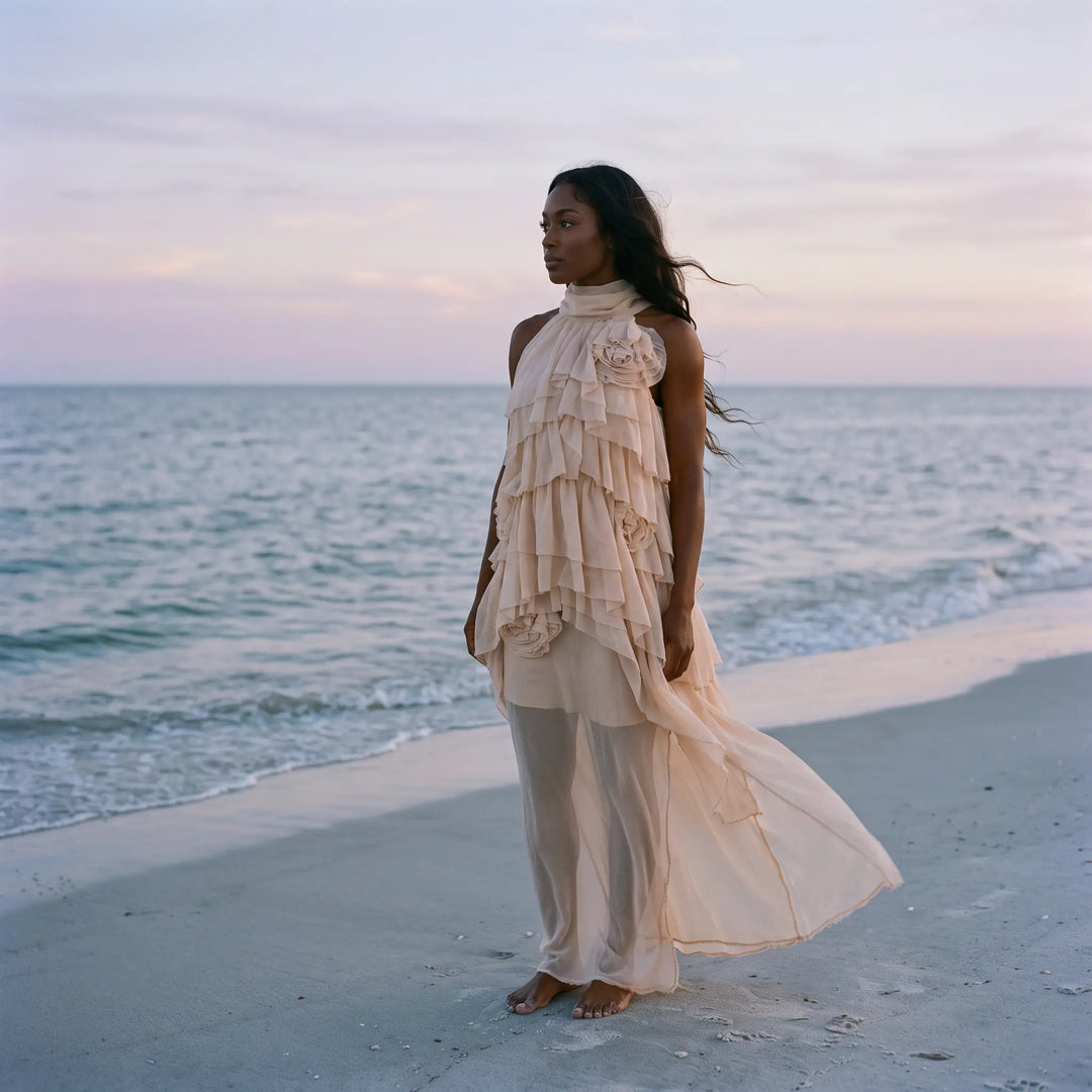 A woman in a flowing, pale nude ethereal maxi dress with layered ruffles stands barefoot on a sandy beach, with the ocean and soft sunset sky in the background.