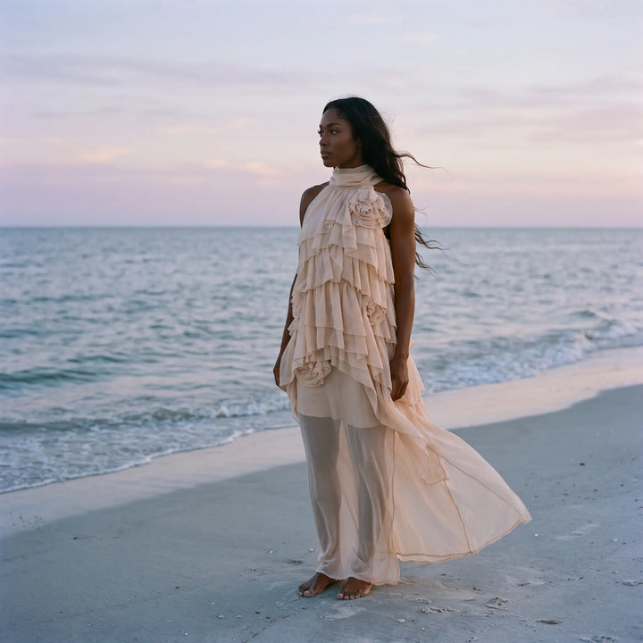 A woman in a flowing, pale nude ethereal maxi dress with layered ruffles stands barefoot on a sandy beach, with the ocean and soft sunset sky in the background.
