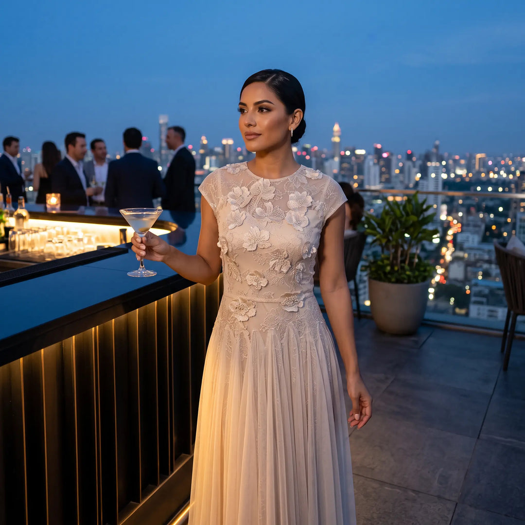 Woman in an elegant blush pink tulle evening gown with delicate floral embroidery on the bodice and cap sleeves, holding a cocktail on a rooftop bar at twilight with city lights in the background.