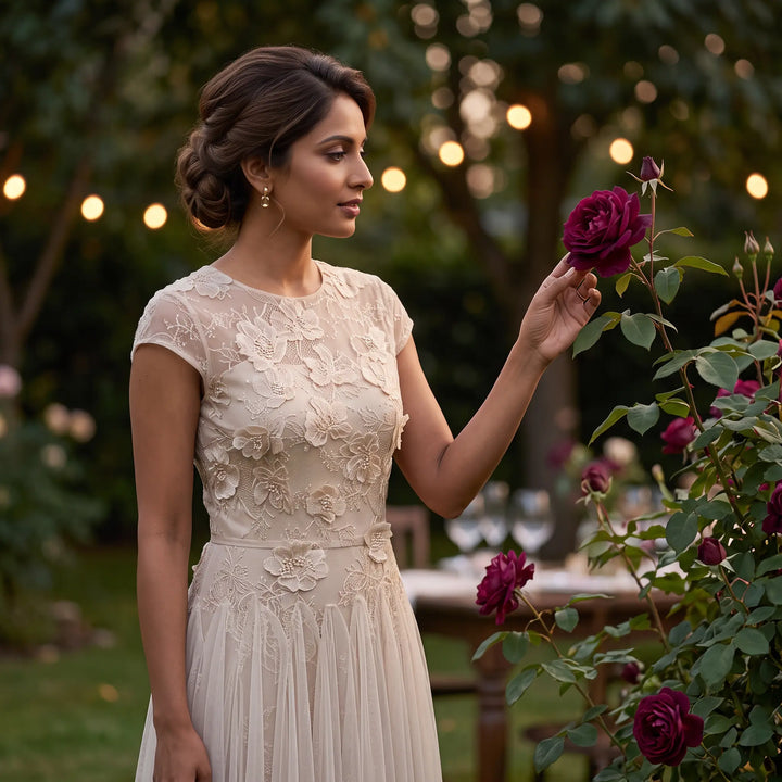 Elegant woman in a floor-length blush pink tulle A-line evening gown with cap sleeves and an intricately embellished lace bodice, standing in a garden setting.