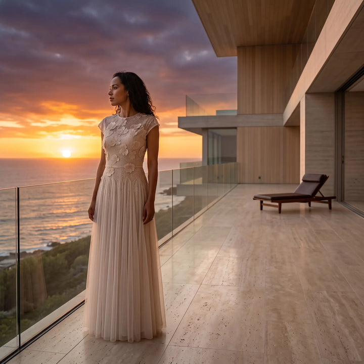 Woman wearing an elegant blush pink tulle A-line evening gown with a detailed lace bodice and cap sleeves, standing on a modern balcony overlooking the ocean at sunset.